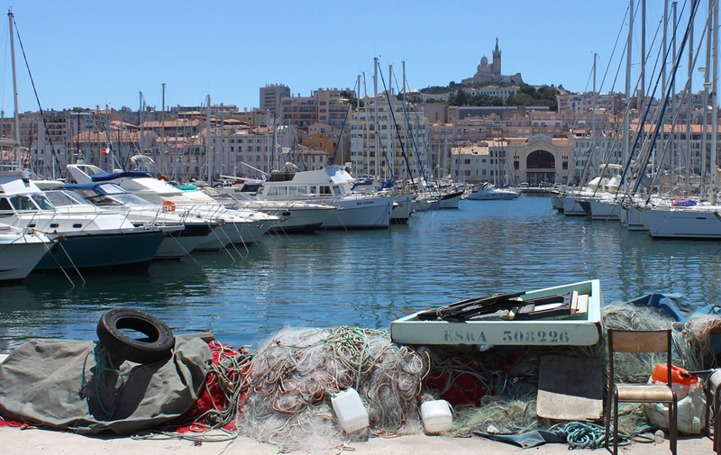 El puerto de Marsella, bordeado de yates elegantes, a lo lejos una estatua en lo alto de una iglesia en una colina...