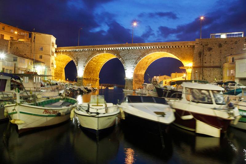 Un puente de piedra que cruza un pequeño puerto de Marsella por la noche, pequeños barcos se alinean a los lados