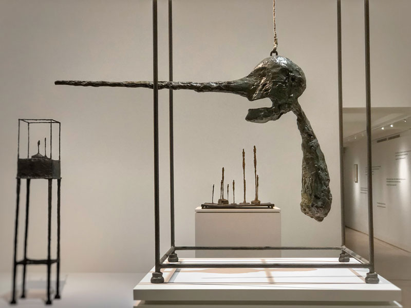 Escultura de Alberto Giacometti, una cabeza redonda y negra con una nariz muy larga y puntiaguda cuelga del techo