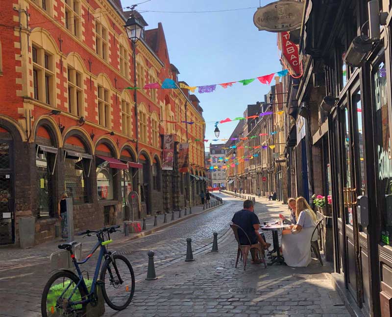 Calle adoquinada en Lille, gente sentada en una cafetería disfrutando del sol y los banderines al otro lado de la calle